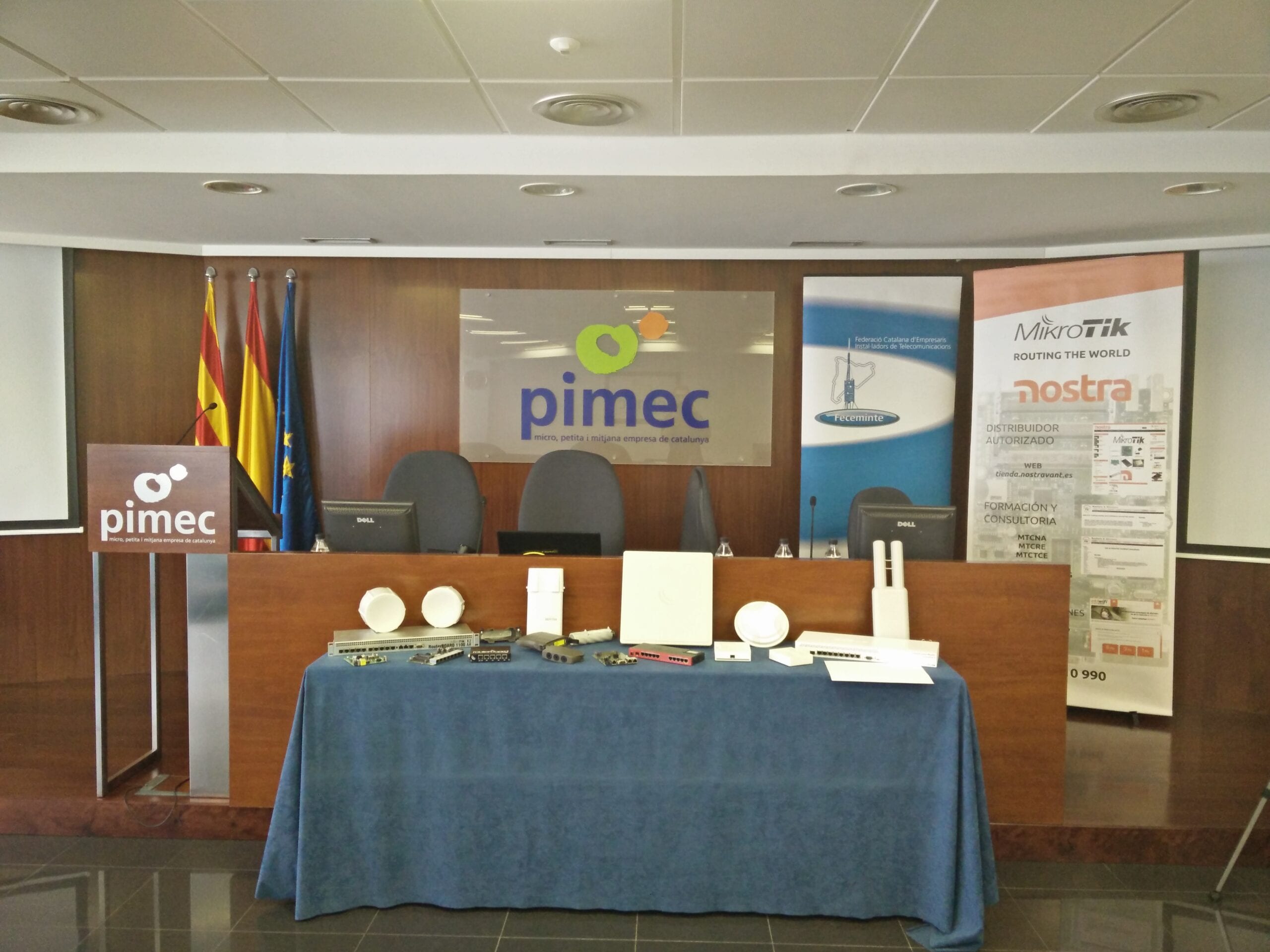 Sala de conferencias de PIMEC con equipos de red MikroTik expuestos sobre la mesa y banners de Feceminte y Nostra
