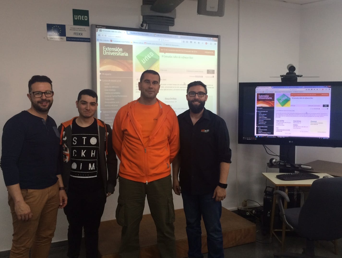 Cuatro personas de pie ante una pantalla de la UNED con la página de las IV Jornadas de software libre