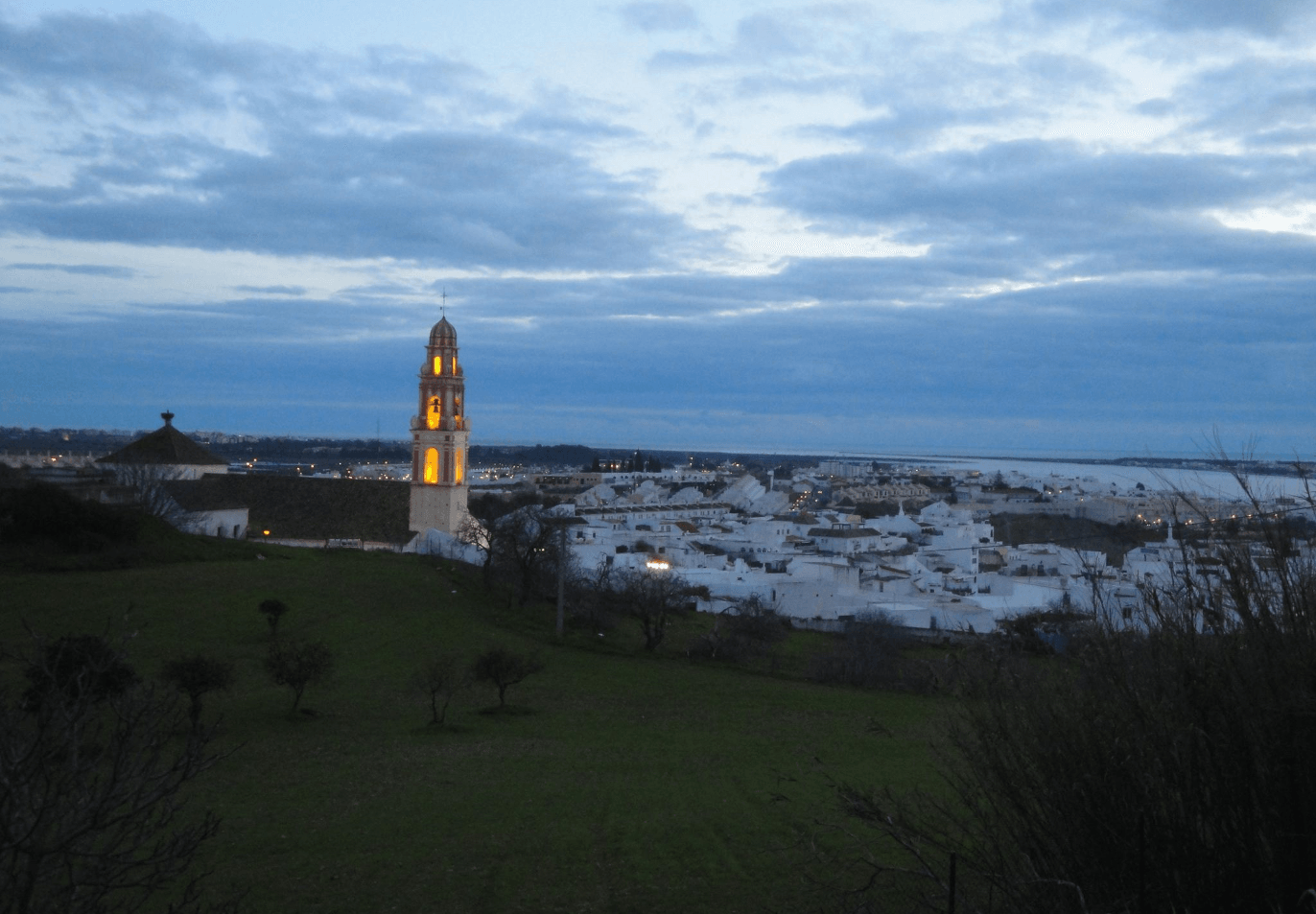 Campanario de una iglesia iluminado al atardecer, con el pueblo blanco y la costa de Huelva al fondo