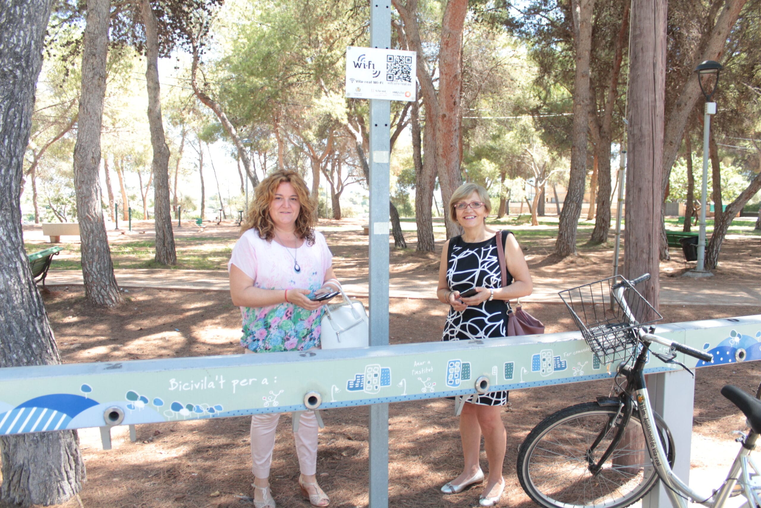 Dos mujeres junto a un cartel de wifi municipal en un parque de Vila-real, con bicicletas de alquiler cerca