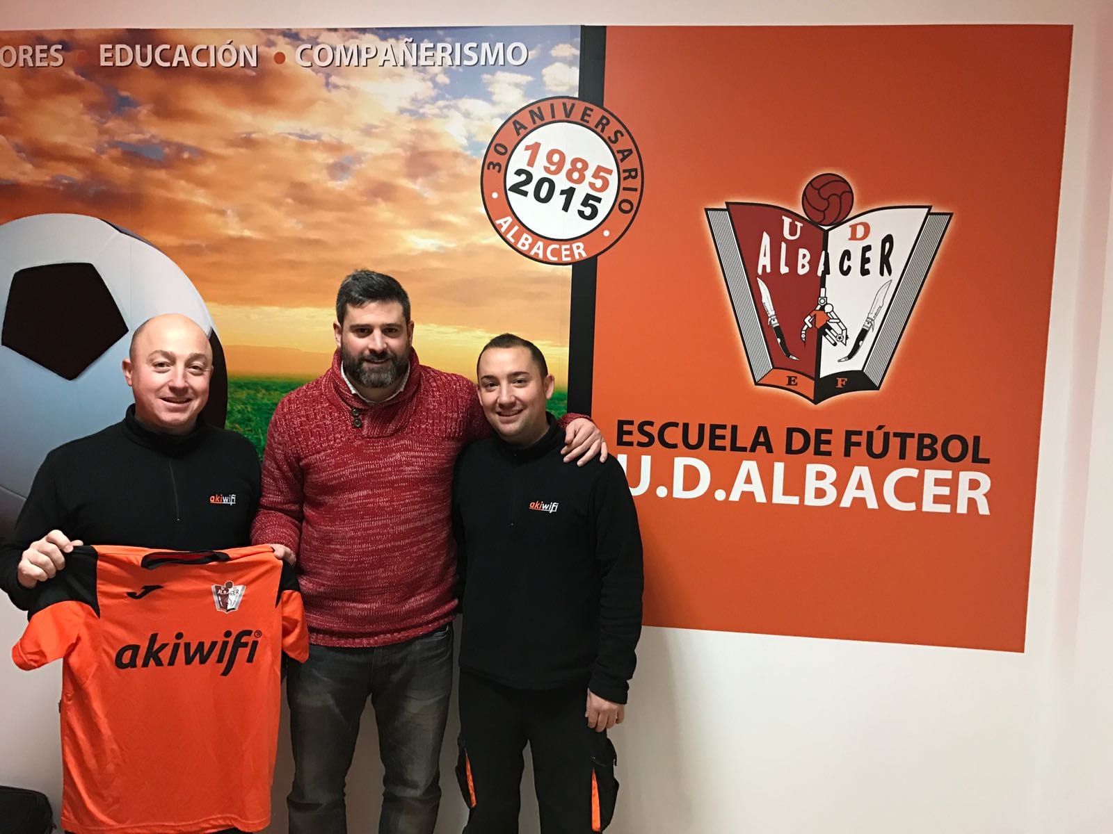 Tres hombres posando con una camiseta patrocinada por akiwifi ante el mural de la Escuela de Fútbol U.D. Albacer