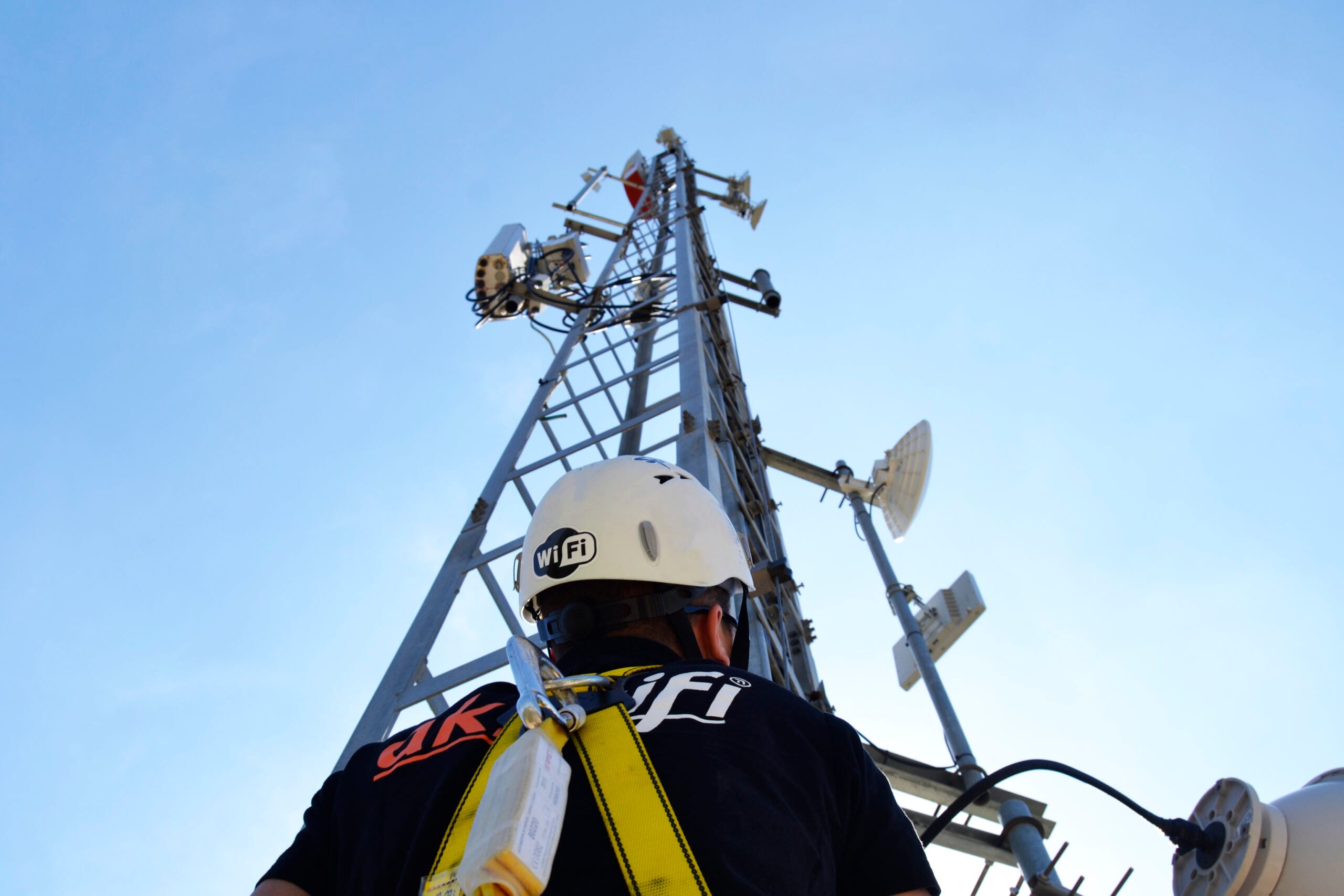 Técnico de akiwifi con casco de obra con el distintivo «WiFi», mirando hacia una torre de telecomunicaciones