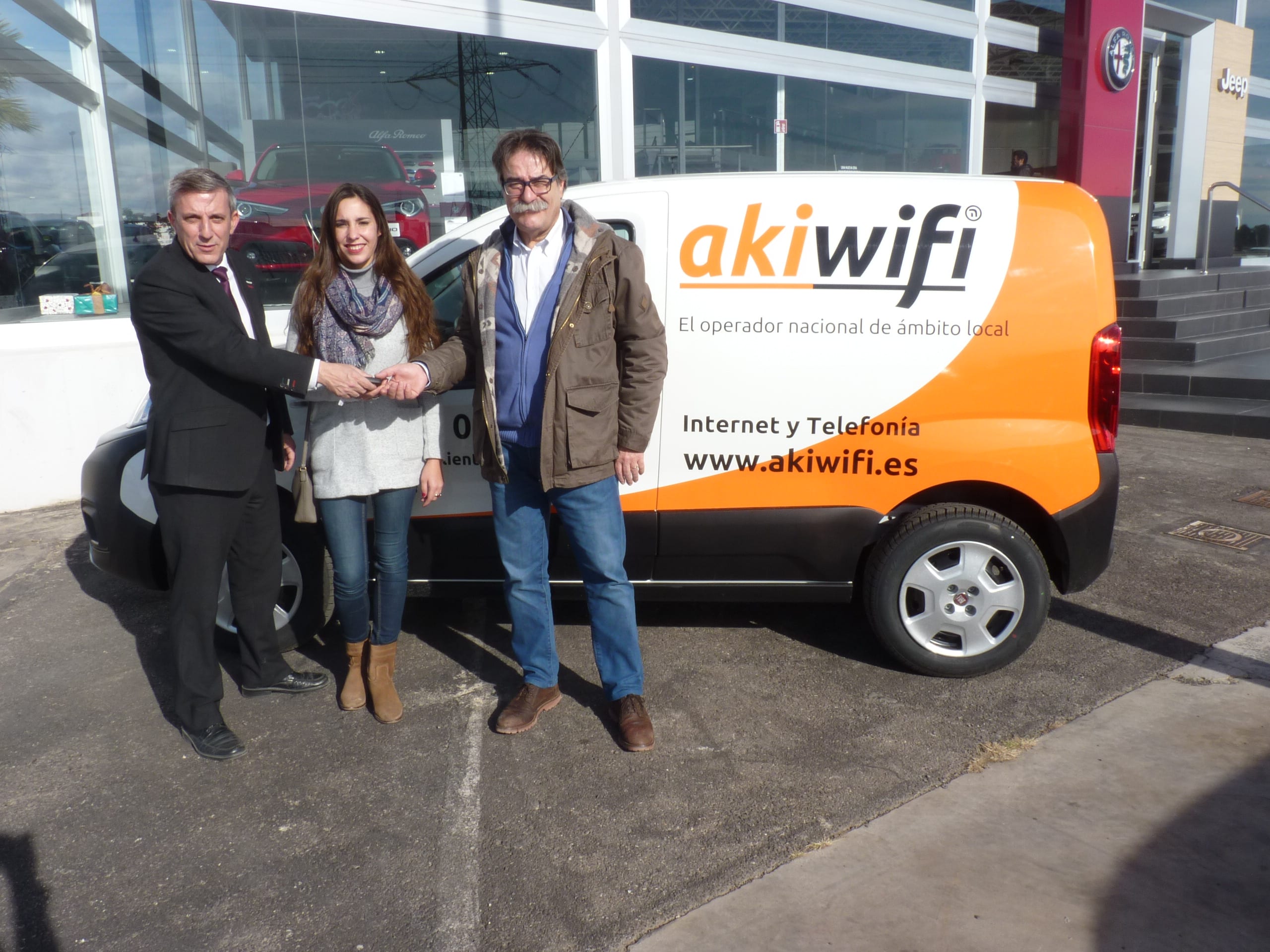 Tres personas entregando las llaves de una furgoneta rotulada con el logo de akiwifi frente a un concesionario