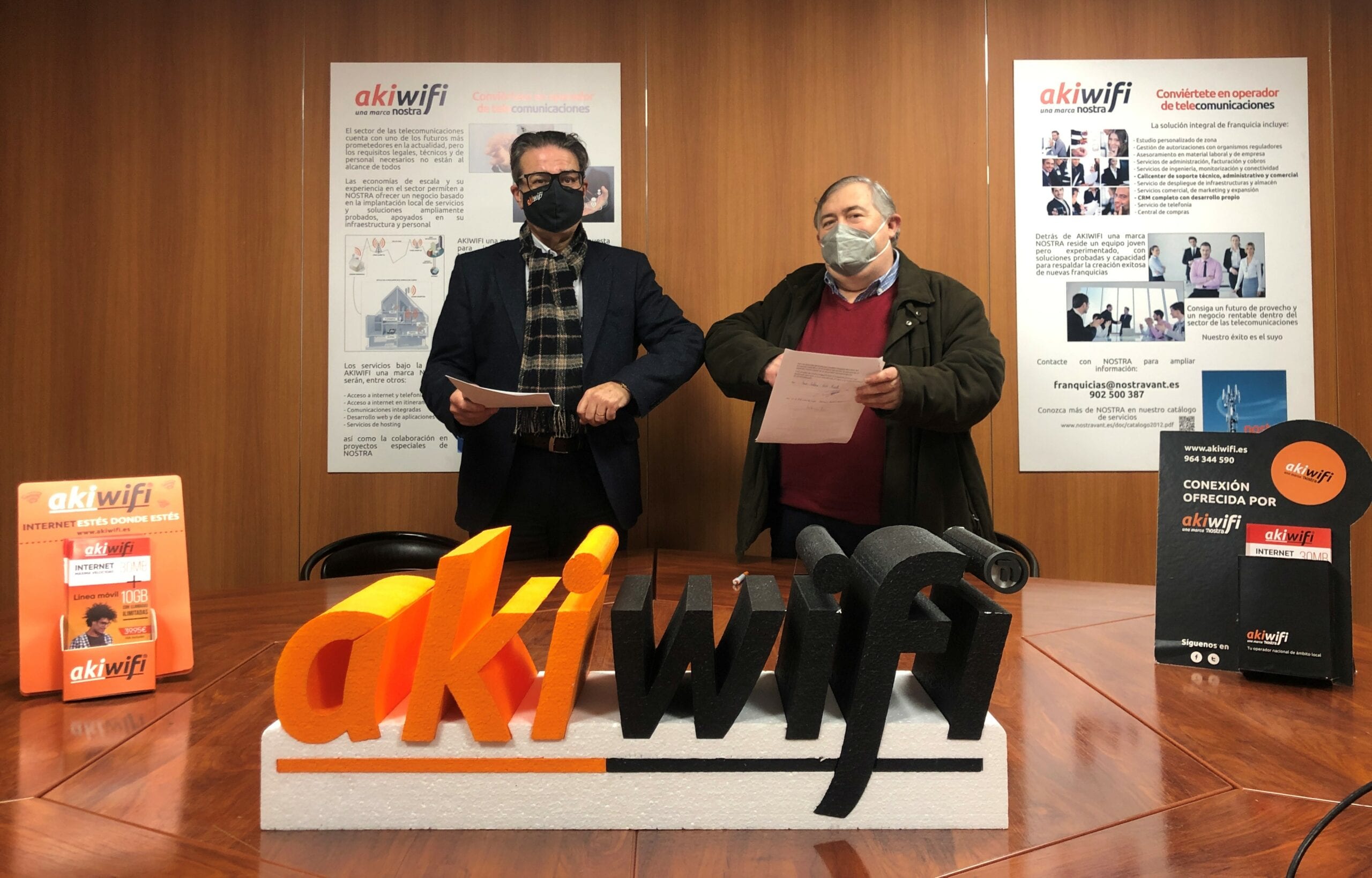 Dos representantes con mascarilla posando junto al logo tridimensional de akiwifi en la firma del acuerdo