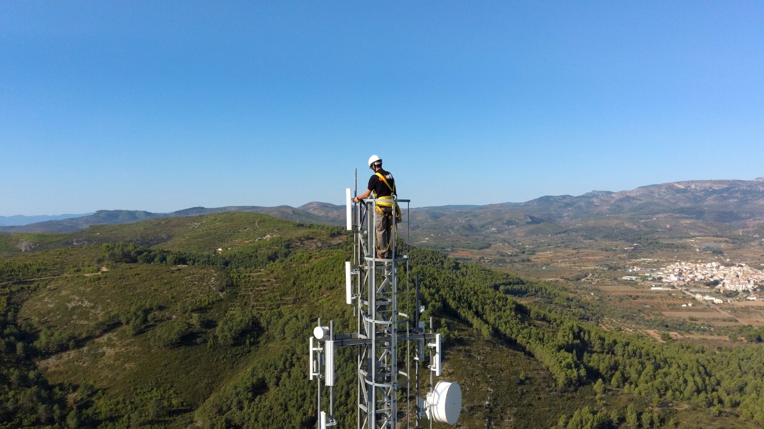 Técnico de akiwifi en lo alto de una torre de telecomunicaciones, con el valle y un pueblo al fondo