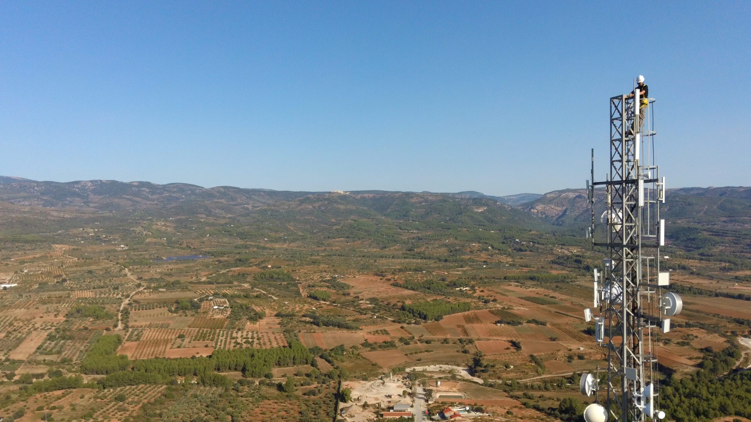 Técnico subido a una antena de telecomunicaciones en lo alto de una colina, con el valle rural al fondo