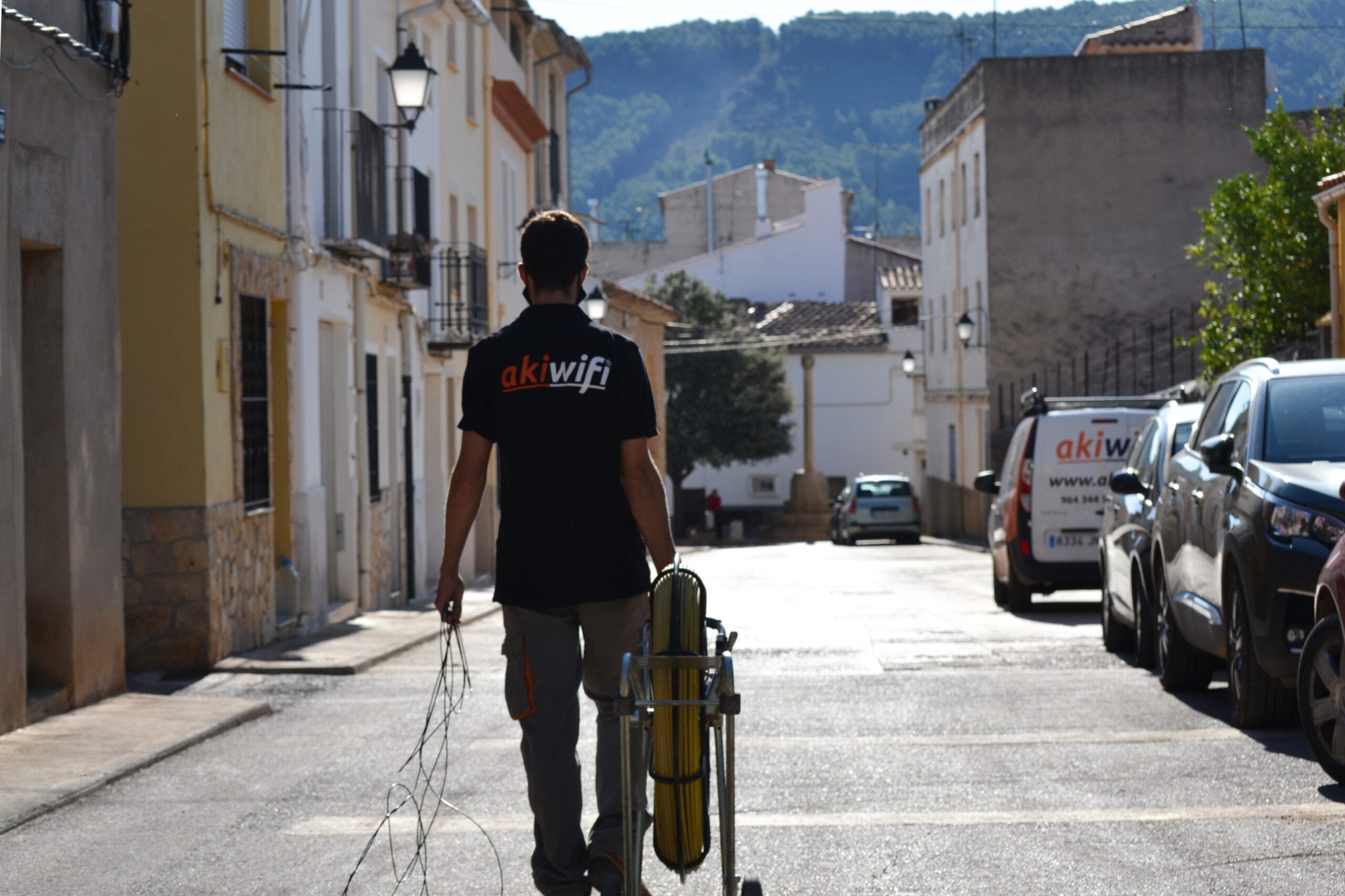 Técnico de Akiwifi caminando por una calle con un carrete de cable de fibra óptica, de espaldas, con la sierra al fondo